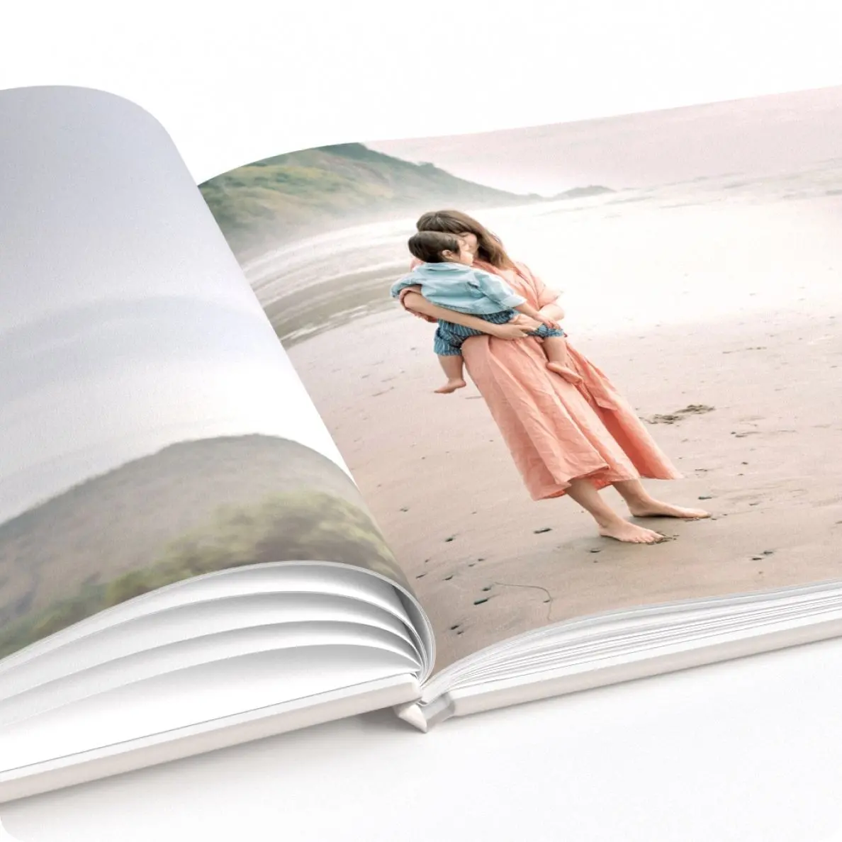 An open photo book on a white surface displaying a page with an image of a woman in a long peach dress holding a child on a sandy beach, evoking a sense of calm and tenderness.
